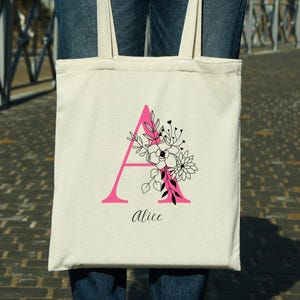 Può includere: Una borsa tote color crema con una grande lettera "A" rosa e un design floreale in bianco e nero. Il nome "Alice" è scritto in corsivo sotto. La borsa ha due manici color crema.