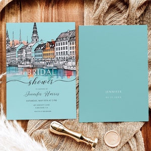 Pode incluir: Um convite de chá de panela azul e branco com uma ilustração em aquarela de uma cidade. O texto diz "BRIDAL shower" em uma fonte de escrita e "Jennifer Harris" em negrito. O convite inclui a data, a hora e o local do evento.