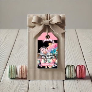 May include: A gift box tied with a beige ribbon, featuring a pink tag with the text "Thanks for coming to my party!" and art supplies. Macarons in green, pink, and grey are placed on either side of the box.