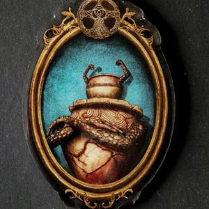 May include: Keychain with an oval pendant showing a detailed illustration of a brown, ornate vessel with a lid, set against a teal background within a gold frame. A Celtic knot design tops the frame.