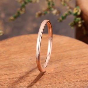 May include: A rose gold wedding band ring. The ring is thin and has a smooth, polished surface. The ring is set against a wooden background with a blurred green plant in the background.