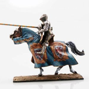 Puede incluir: Figura detallada de un caballero con armadura plateada montado a caballo. El caballo está cubierto con una tela azul y dorada con escudos heráldicos. El caballero sostiene una larga lanza con rayas naranjas y azules alternas.