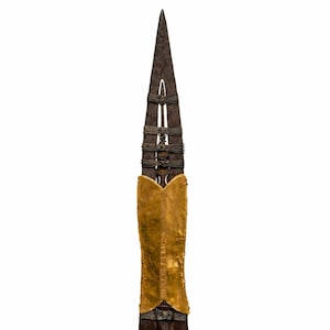 Puede incluir: Una lanza con una hoja oscura y puntiaguda y un asta de madera. El asta está envuelta con metal y cuerda, y presenta un elemento dorado en forma de escudo. La lanza está sobre un fondo blanco.