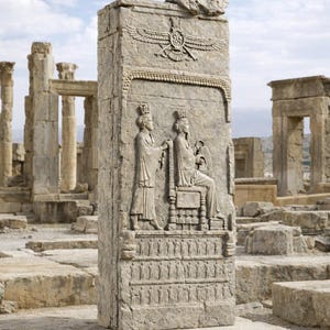 May include: A weathered stone pillar with carved relief depicting figures in ancient attire. The pillar stands amidst ruins under a cloudy sky. The carvings include a winged symbol and figures in regal attire.