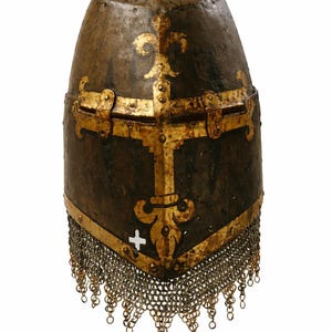 May include: A medieval helmet with a dark, aged appearance. It features gold-coloured decorative elements, including a cross and fleur-de-lis design. A chainmail fringe hangs from the bottom. The helmet is likely made of metal.