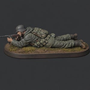 May include: A miniature figure of a WWII German soldier in a green and grey uniform, aiming a submachine gun while lying down. The figure wears a helmet and carries a backpack. The base is oval, with grass. The figure is a detailed model.