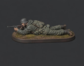 Puede incluir: Figura en miniatura detallada de un soldado alemán de la Segunda Guerra Mundial en uniforme, tumbado y apuntando con una ametralladora. La figura está pintada en tonos verdes y grises, con casco y mochila. La base es ovalada y con hierba.