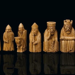 May include: A collection of intricately carved chess pieces, each with a unique design and expression. The pieces are a light beige color and stand against a black background. The set includes a shield, a knight on a horse, a bishop, a king, and a queen.