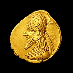 May include: A gold coin featuring a profile of a man with a beard, wearing a detailed headdress and ornate clothing. The coin has a rough, irregular edge and is set against a black background.