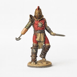 May include: A miniature gladiator figurine in a dynamic pose, clad in red and bronze armor. The figure holds a curved blade in one hand and a short sword in the other. The helmet has a red crest, and the figure stands on a small, round base.
