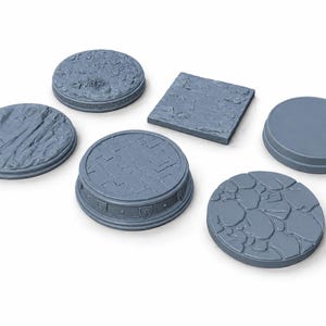May include: A collection of six gray miniature bases in various shapes and textures. The set includes circular and square bases featuring detailed surfaces such as cobblestone, cracked earth, rocky terrain, and smooth flat tops with shield motifs.