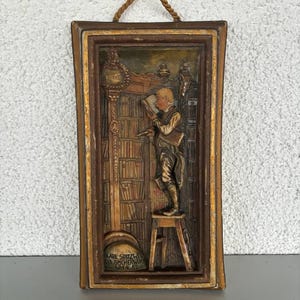 May include: A rectangular, framed relief sculpture depicting a person reading in a library. The artwork features a person standing on a stool, reading a book in front of a bookshelf. The frame is brown with gold accents, and a rope is attached for hanging.