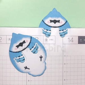 May include: Two bluebird shaped paper bookmarks with white accents and black eyes. The bookmarks are placed on a page of a planner with a grid pattern.