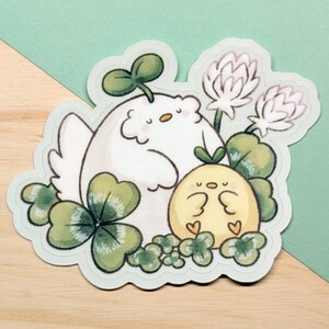 May include: A die-cut sticker featuring two cartoon chicks and clovers. One chick is white with a green sprout on its head, the other is yellow. The sticker has a light green border and is set against a wooden surface and a teal background.
