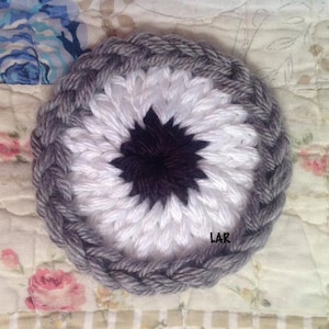 May include: A crocheted coaster with a white center, a black starburst design, and a gray outer ring. The coaster is on a floral fabric background.