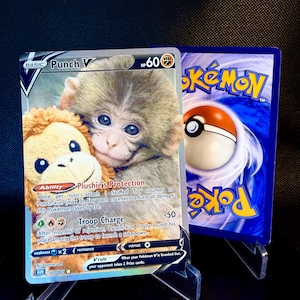 May include: A Pokémon trading card, "Punch V", displaying a monkey and a plush toy. The card has a HP of 60 and text detailing abilities and attacks. The reverse side shows a Poké Ball design on a blue background, with the word "Pokémon".