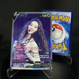 May include: A holographic Pokémon trading card featuring "Ella V" with a portrait of a smiling woman. The card has a purple background and includes text such as "Natural Spotlight" and "Butterfly Bloom". A second Pokémon card is visible in the background.