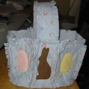 以下が含まれることがあります： フリルのある青い布製のイースターバスケット。バスケットには3つの仕切りがあり、それぞれに異なるアップリケが施されています。ピンクの卵、茶色のウサギ、黄色の卵です。