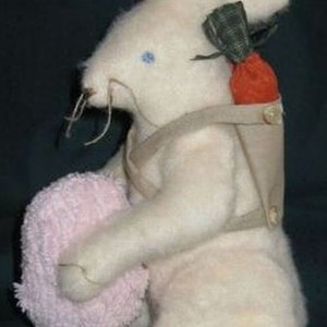 Puede incluir: Un juguete de peluche de conejo blanco con un arnés de tela marrón y que lleva una toalla rosa y una pequeña zanahoria naranja en una bolsa de tela verde.
