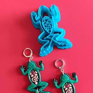May include: A pair of green frog earrings with glitter, each with a detailed anatomical design of the frog's internal organs, including a heart, lungs, and intestines. Each frog has three silver daggers hanging from its legs.