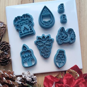 May include: A set of eight blue plastic cookie cutters featuring holiday-themed shapes including a fireplace, a teardrop with a brick pattern, two small boots, a dove, a candle with a face, a pinecone, and a light bulb with a bow design.