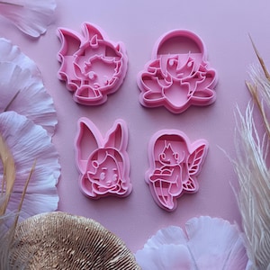 May include: A set of four light pink plastic cookie cutters featuring stylized character designs. The shapes include a horned figure, a character with large ears, a figure with long hair and wings, and a seated character with wings.