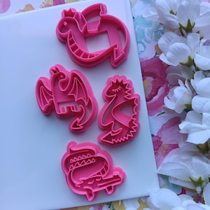 May include: Four pink plastic cookie cutters in dragon shapes. The cutters include a dragon with wings, a dragon with a crown, and a dragon with a long neck. The cutters are on a white surface.