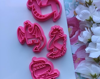 May include: Four pink plastic cookie cutters in dragon shapes. The cutters include a dragon with wings, a dragon with a crown, and a dragon with a long neck. The cutters are on a white surface.