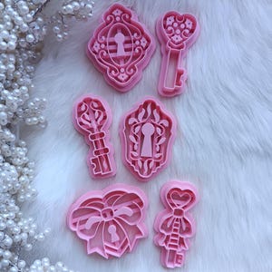 May include: Pink cookie cutters in various shapes, including keys, keyholes, and a bow. The cutters are arranged on a white, fluffy surface, with pearl-like decorations in the background. The designs are detailed and suitable for baking or crafting.
