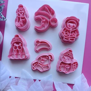 May include: A set of seven pink plastic cookie cutters featuring various winter-themed shapes, including a snowman, a crescent moon with a hat, a bear in a parka, a walrus in a beanie, a small hat, a pig in a scarf, and a beaver in a hat.