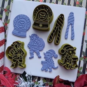 May include: A collection of light purple cookie cutters and their corresponding stamped impressions featuring a yeti in a snow globe, a wolf-like character in a Santa suit, a small creature with a stocking, and a pair of striped candy cane shapes.