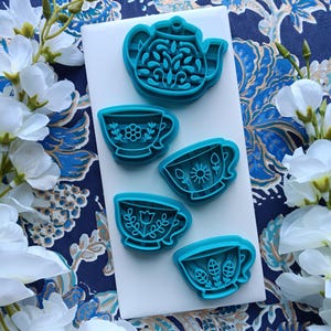 May include: A collection of teal-colored cookie cutters in the shape of a teapot and teacups. The teacups feature floral and leaf designs. The cutters are arranged on a white surface, with a blue floral patterned background.