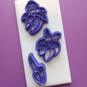 May include: Three purple plastic cookie cutters featuring distinct designs: one shaped like a banana with a face, one shaped like a banana with a lipstick tube, and one shaped like a single banana. The cutters are arranged on a white rectangular base.