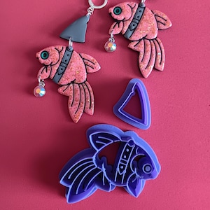 May include: A pair of pink glitter fish earrings with black accents and a silver hoop. The earrings are hanging from a gray triangle charm. A purple fish-shaped cookie cutter is also included.