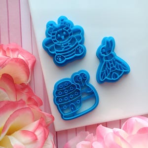 May include: Three blue plastic cookie cutters shaped like a bee, a snail, and a ladybug. The bee has a striped body and wings. The snail has a shell with a door and stripes. The ladybug has spots and wings.
