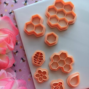 May include: A set of eight orange plastic cookie cutters shaped like honeycombs, a bee, a flower, a honeycomb, and a teardrop. The cutters are on a white surface.