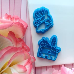 May include: Two blue plastic cookie cutters, one shaped like a cow with wings and the other shaped like a butterfly. The cutters are on a white surface with pink and white striped background.