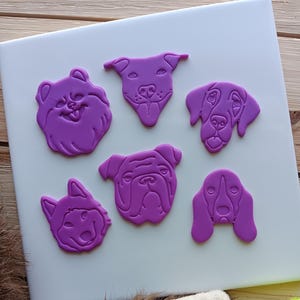 May include: Six purple plastic dog breed cookie cutters on a white surface. The cutters are shaped like the heads of a bulldog, a husky, a beagle, a labrador, a pit bull, and a pomeranian.
