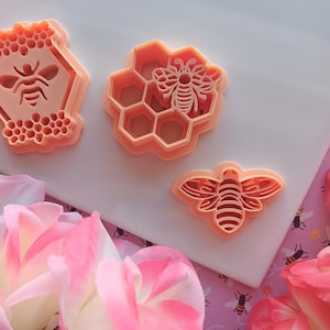 May include: Three peach-colored cookie cutters shaped like honeycombs and bees. The cutters are on a white surface with pink flowers in the background.