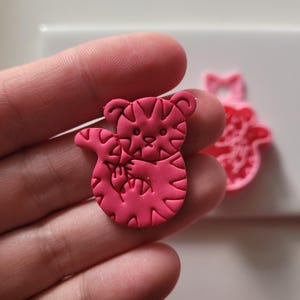 May include: A vibrant pink tiger-shaped polymer clay charm with detailed stripes and facial features. The charm is held in a hand, with a second, similar charm visible in the background. The tiger charm is a unique, handmade item.