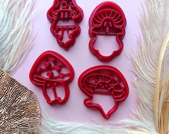 May include: Four red plastic mushroom cookie cutters on a pink background. The cutters are different sizes and shapes, with some featuring details like dots and lines.