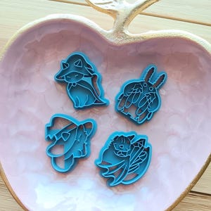 May include: Four blue plastic cookie cutters in various fantasy creature shapes, including a fox, a rabbit, and a dragon. The cutters are arranged in a pink apple-shaped dish with a gold leaf accent.