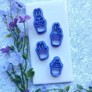 May include: Four blue plastic cookie cutters in the shapes of Easter bunnies and a cupcake. The cutters are on a white surface, with a blue floral patterned background and purple flowers.