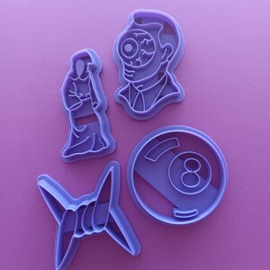 May include: Set of four purple plastic cookie cutters. The cutters are shaped like a man with one eye, a 8-ball, a shuriken, and a person with a long neck.