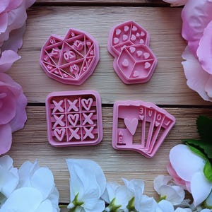 May include: Four pink cookie cutters in various shapes: a heart with numbers, two dice with heart-shaped pips, a tic-tac-toe board with hearts, and a playing card hand with the word "LOVE". The cutters are surrounded by pink and white flowers.