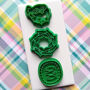 May include: Three green cookie cutters in various shapes: a monster face, a wreath of fish, and a circular design with three fish. The cutters are on a white surface, set against a colorful plaid background.