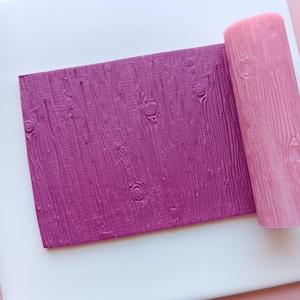 May include: A purple textured rolling pin and a matching rectangular sheet with a wood grain pattern. The rolling pin is cylindrical and pink. The sheet and rolling pin are on a white surface, with a pink background.