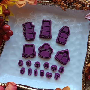 May include: A collection of purple cookie cutters in various shapes, including a house, a person, and mushrooms, arranged on a white surface. The cutters are made of plastic and are designed for baking. The image is taken from a high angle.