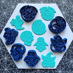 May include: A collection of dark blue and bright turquoise plastic cookie cutters featuring Halloween-themed designs including a bat, a ghost, a cat, a skull, a vampire, and a witch. The set includes both outline cutters and detailed stamps.