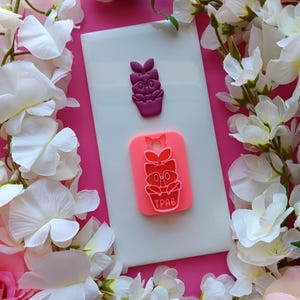 May include: Two silicone molds, one purple and one pink, featuring a cartoon flower pot design. The purple mold is a single flower pot, while the pink mold has the text "TPAB" at the bottom. The molds are on a white surface, surrounded by white and pink flowers.